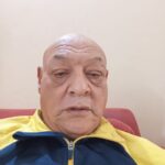 Retrato de Vicente Sampaio olhando para a câmera. Ele veste uma jaqueta esportiva azul e amarela e está sentado em um ambiente interno de parede clara, com expressão serena